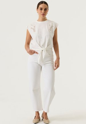 Frau trägt ein ärmelloses weißes Oberteil mit Knoten vorne und Ösen-Details, kombiniert mit hochtaillierten weißen weit geschnittenen Jeans und beigen flachen Schuhen.