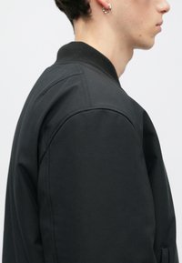 Melna bumbieru jaka ar gludu tekstūru, ribotu apkakli un pastiprinātiem šuvēm uz pleciem. Aprīkota ar sānu kabatu.