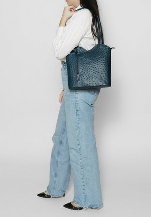 Bucketstijl tas van donkerblauw textuurmateriaal, met een ritsluiting, een gebogen bovenkant en een enkele handgreep. Draagt bij lichtblauwe jeans.