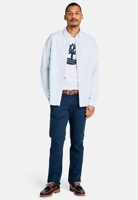 Chemise bleu clair à manches longues, t-shirt blanc avec motif en dessous, pantalon bleu marine, chaussures marron à lacets et ceinture marron. Design simple et coupe décontractée.