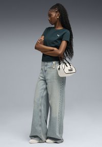 Camiseta verde oscura con un pequeño logo blanco, combinada con jeans de pierna ancha azul claro. Bolso blanco con acentos y logo negros, llevado de manera informal.