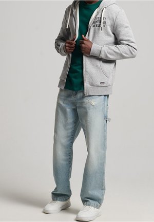 Felpa grigia con zip sopra una maglietta verde, abbinata a jeans di denim blu chiaro con effetto usurato e scarpe da ginnastica bianche. Design semplice e casual.