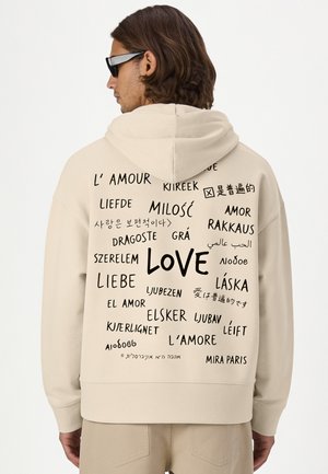 Personne portant un sweat à capuche beige avec le mot "love" écrit en plusieurs langues dans le dos, dos à la caméra.