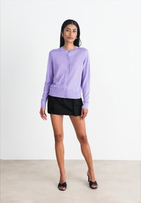 Lila cardigan i mjukt stickat material med rund halsringning och knappar framtill, matchad med en svart minikjol och svarta öppna skor.