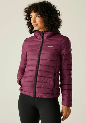 Woman with curly hair wearing a zipped purple puffer jacket with hood and black leggings, standing with hands in pockets.