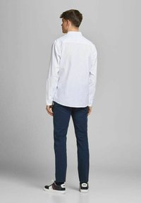 Camicia bianca a maniche lunghe, vista posteriore, abbinata a pantaloni blu navy e sneakers nere. Materiale in cotone con vestibilità casual, bottoni e colletto visibili.