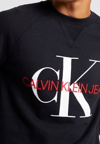 Felpa nera con grandi lettere bianche "CK" e logo ricamato rosso "Calvin Klein Jeans" sulla parte anteriore indossata da una persona.