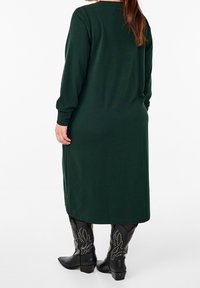 Langes, dunkelgrünes Kleid mit lockerer Passform, vollem Ärmel und geradem Schnitt; kombiniert mit schwarzen, mit Nieten verzierten Westernstiefeln.