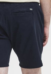 Mørkeblå shorts med en glatt tekstur, som har en baklomme, elastisk linning og et lite merke sydd bakpå.