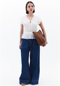 Blusa bianca a trama con maniche corte e scollatura legata; abbinata a jeans azzurri a gamba larga; tiene in mano una borsa intrecciata marrone.