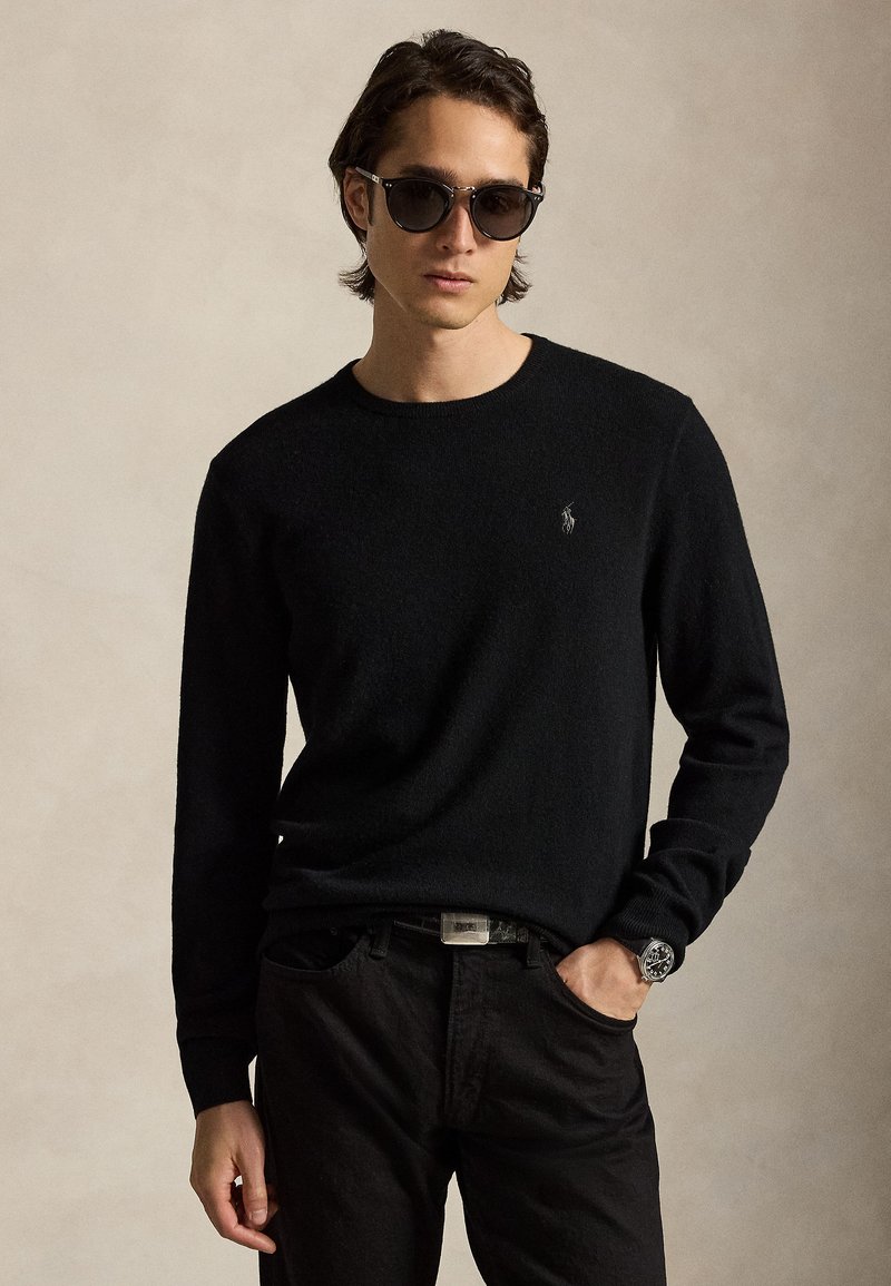 Junger Mann, der einen schwarzen Pullover, schwarze Hose, Sonnenbrille und Uhr trägt, steht mit einer Hand in der Tasche vor einem einfarbigen Hintergrund.