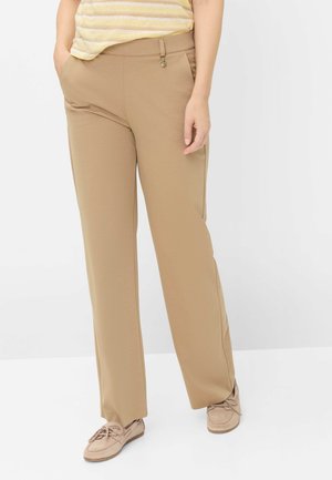 Person iført beige bukser med lige ben og lommer samt beige snørestøvler, vist fra taljen til fødderne på hvid baggrund.