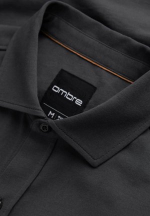 Polo shirt gris foncé avec un col pointu, une patte de boutonnage et une étiquette portant le mot "ombre". Texture douce et lisse, design classique.