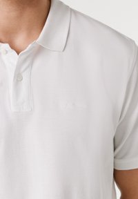 Ασπρό πόλο μπλουζάκι από υφασμάτινη υφή, με δύο κουμπιά στη λαιμόκοψη και διακριτικό κεντημένο λογότυπο στην αριστερή πλευρά του στήθους. Κοντά μανίκια.