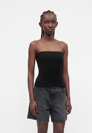 Kvinde med flettet hår iført en sort stropløs top og løse mørke denimshorts, der står mod en ensartet lys baggrund.