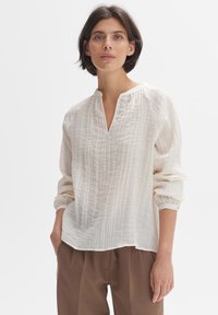 Blusa leggera in crema con scollatura a V e maniche lunghe a sbuffo. Tessuto testurizzato con sottili strisce verticali. Abbinata a pantaloni marroni.