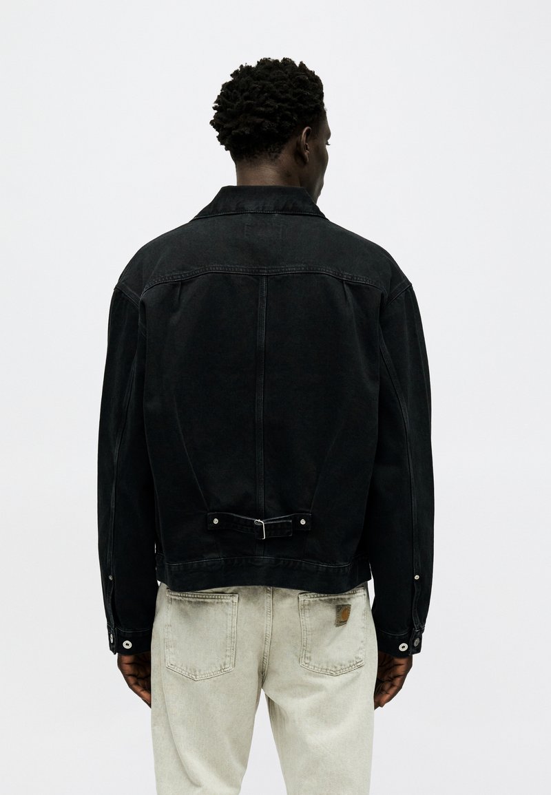 Homme aux cheveux courts et bouclés portant une veste en denim noire et un jean beige clair, vu de dos sur un fond uni.