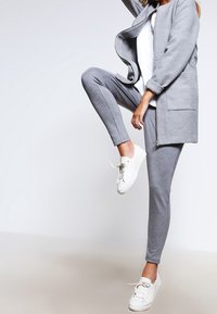 Tuta in cotone grigia con giacca con zip e pantaloni affusolati, indossata con sneakers bianche. Il tessuto appare morbido e liscio.