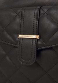 Zwarte, gewatteerde leren tas in close-up, met een gestikte riem en een gouden sluiting gegraveerd met "ANNA FIELD".