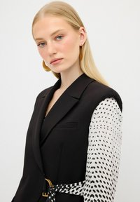 Blazer noir au toucher lisse, avec une manche blanche à motif et une taille ceinturée. Des boucles d'oreilles en or en forme de cercle complètent le look.
