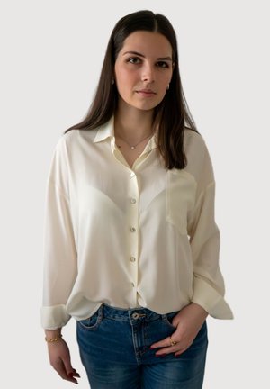 Jeune femme aux longs cheveux bruns portant un chemisier crème à boutons et un jean bleu, debout devant un fond clair uni.