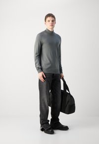 Harmaa poolopaita, slim fit, paritettu tummiin farkkuihin. Malli pitää mustaa laukkua. Silkkinen neulostekstuuri, minimaalinen brändäys, rento muotoilu.