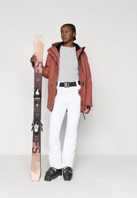 Síruha rozsdavörös dzsekivel, szürke felsővel, fehér nadrággal, fekete övvel és sícipőkkel. Síléceket tart kötésekkel, szürke háttéren.