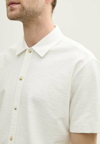 Korte mouwen witte shirt van een textuurstof, met een puntige kraag en lichtgekleurde knoopsluitingen aan de voorkant.