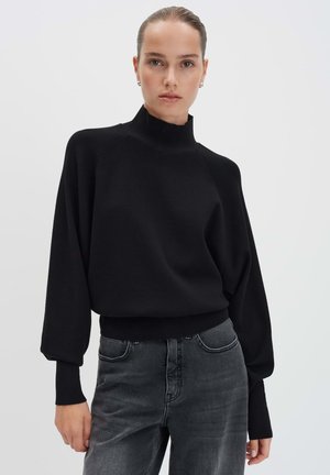Frau mit zurückgegeltem Haar, die einen schwarzen Pullover mit hohem Kragen und Ballonärmeln sowie ausgewaschenen schwarzen Jeans vor einem schlichten Hintergrund trägt.