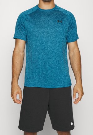 Mann trägt ein türkisfarbenes kurzärmeliges Under Armour-Shirt und schwarze Nike-Shorts und steht mit entspannten Armen an den Seiten.