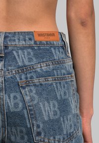 High-waisted denim jeans in light blue with a subtle all-over logo pattern, featuring orange leather patch and visible stitching details.