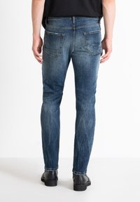Jeans de mezclilla azul con un ajuste ajustado, pierna recta y lavado desgastado, con dos bolsillos traseros y detalles de costura marrón. Se ven zapatos negros.