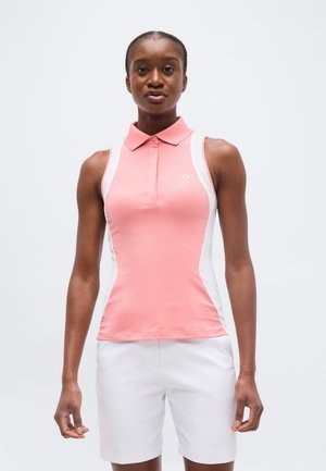 Frau trägt ein ärmelloses rosa-weißes Hemd mit Kragen und weiße Shorts, steht vor einem einfarbigen hellen Hintergrund.