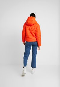 Person klädd i en ljus orange huva pufferjacka, blå jeans och vita sneakers som står mot en enkel vit bakgrund.