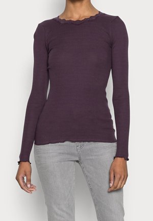 Femme portant un haut moulant côtelé à manches longues violet foncé avec des bords festonnés, assorti à un jean gris clair.