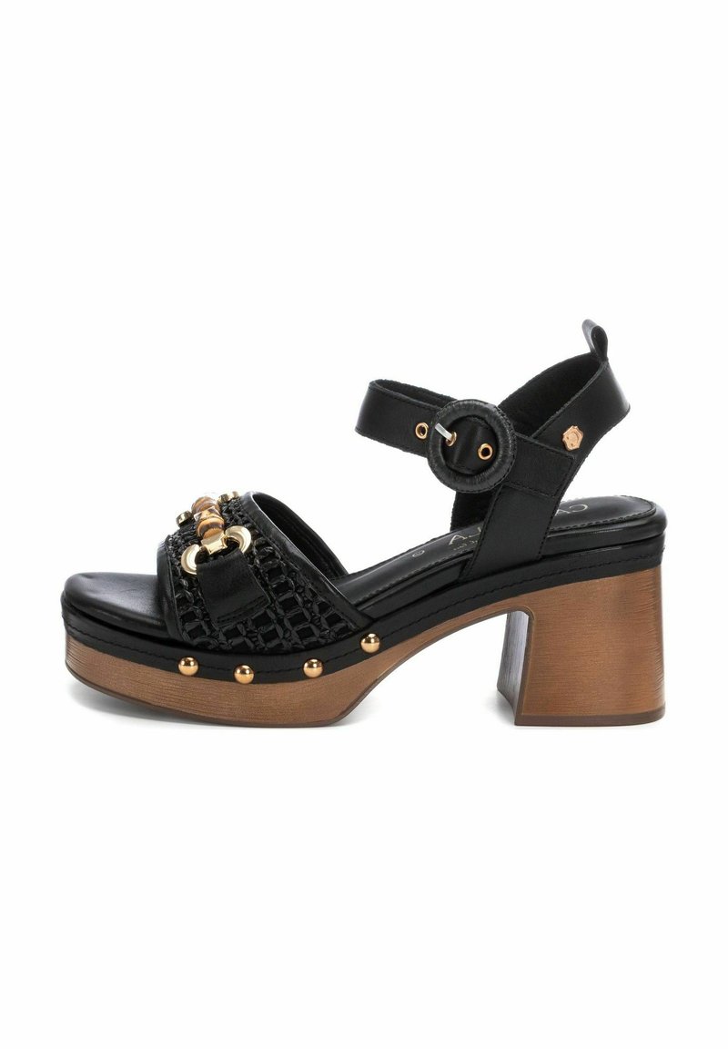 Sandalias de tacón negro con textura tejida, un tacón ancho y cuadrado, tachuelas doradas, una correa para el tobillo ajustable y herrajes decorativos dorados.