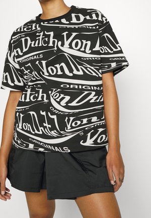 Henkilö pukeutunut mustaan lyhythihaiseen paitaan, jossa on suuret valkoiset "Von Dutch Originals" logot, ja mustiin shortseihin valkoista taustaa vasten.