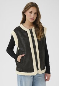 Mlada žena s dugom kosom koja nosi crno-bijeli jaknu od ovčje kože bez rukava preko crnog dugog rukava i svijetlopto blue trapericama.