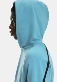 Ung person iførende en lyseblå hoodie med hætten oppe, vist i profil mod en ensfarvet hvid baggrund.
