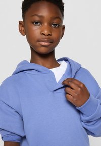 Felpa blu chiaro con una texture morbida, dotata di tasca a marsupio e uno strato interno bianco, posata sulla spalla di un bambino con la pelle scura.