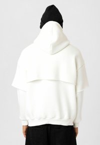 Witte oversized hoodie met een gelaagd ontwerp, voorzien van een capuchon en ribkorte mouwen. De stof heeft een soepele textuur, van achteren getoond.