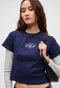 Tumši zila kokvilnas īsa t-krekla ar īsām piedurknēm un apaļu kaklu, uz krūtīm ar baltu tekstu "JOIN THE Club".