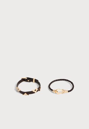 Deux bracelets en cuir noir sur une surface blanche, l'un avec une boucle dorée et des accents décoratifs, l'autre tressé avec une fermeture dorée.