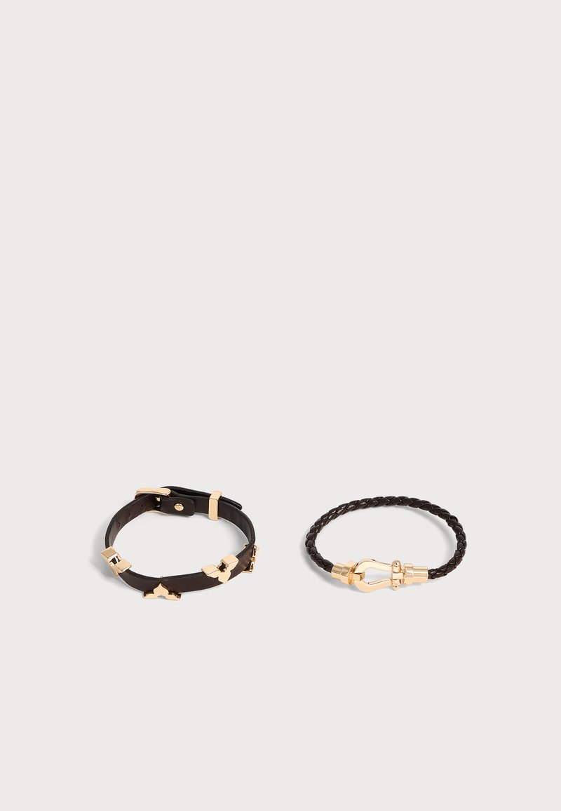 Deux bracelets en cuir noir sur une surface blanche, l'un avec une boucle dorée et des accents décoratifs, l'autre tressé avec une fermeture dorée.