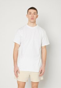 T-shirt en coton blanc à manches courtes et col rond, porté par une personne se tenant contre un fond uni, associé à un short beige.