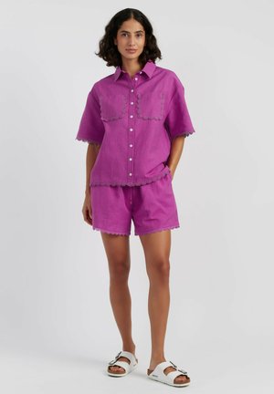 Donna con capelli scuri e mossi che indossa una camicia viola a maniche corte con bottoni e pantaloncini coordinati con bordo in pizzo, sandali bianchi, in piedi con una mano in tasca.