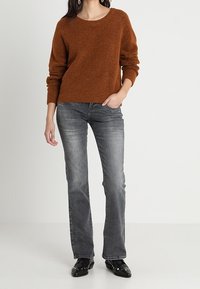 Suéter de punto marrón texturizado con cuello redondo, combinado con vaqueros de campana grises. Botines negros con detalles en tachuelas plateadas.