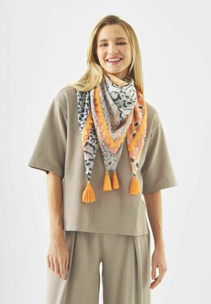 Une femme souriante porte une chemise et un pantalon amples de couleur taupe, accessoirisés d'un foulard triangulaire multicolore orné de pompons orange et de motifs.