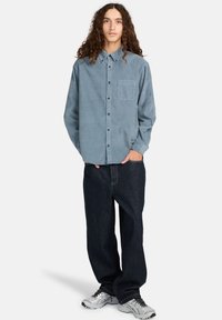 Lyseblå, knappet skjorte med brystlomme og lange ærmer, parret med mørkeblå, løstsiddende jeans og sølvfarvede sneakers.