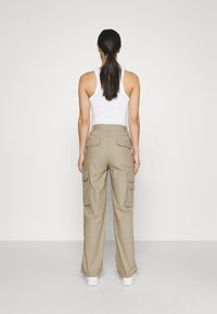 Beige cargobroek met wijde pijpen, voorzien van meerdere zijzakken en een hoge taille. Het model draagt een witte tanktop en sneakers.